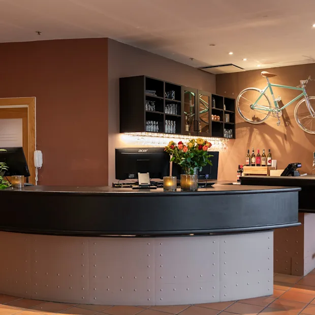 A stylish hotel reception and bar area with a curved counter, monitors, flowers, and a bicycle mounted on a brown wall.