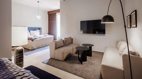 Modern living room with a beige sofa, adjacent bedroom area, art on walls, and a large floor lamp, illustrating a sleek interior.