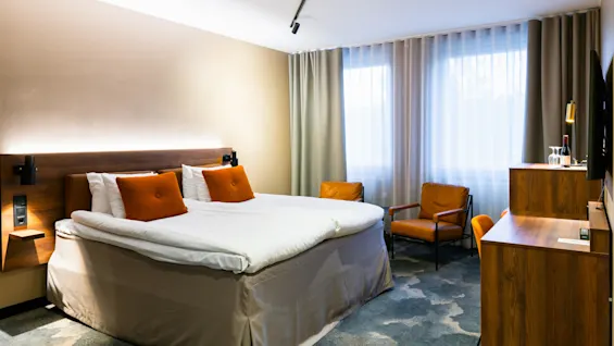 En stor seng med orange puder står centralt i et hotelværelse. To lænestole og et skrivebord i træ er placeret nær store gardinerede vinduer.