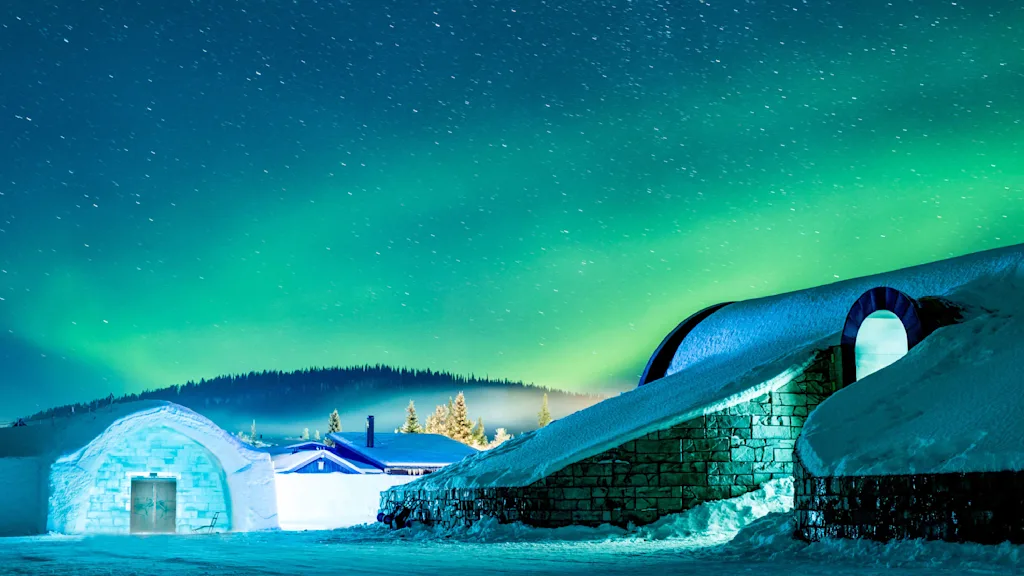 Snedækkede bygninger lyser op under et nordlys på en vinternat.