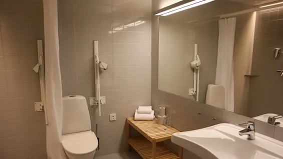 Et toalett står ved siden av en trehylle med sammenlagte håndklær og en toalettrull, alt i et flislagt bad med stort speil og moderne vask.
