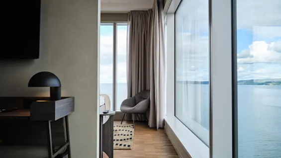 Et værelse med en hyggelig stol nær gulv-til-loft-vinduer, der har udsigt over et roligt hav og blå himmel, med et skrivebord i træ og blød belysning.