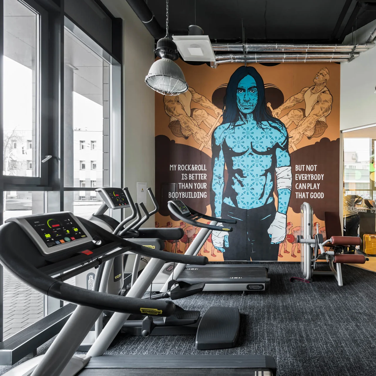 Löpband står stilla i ett gym, vända mot en väggmålning av en blåhyad figur. Texten lyder: "MY ROCK&ROLL IS BETTER THAN YOUR BODYBUILDING BUT NOT EVERYBODY CAN PLAY THAT GOOD."