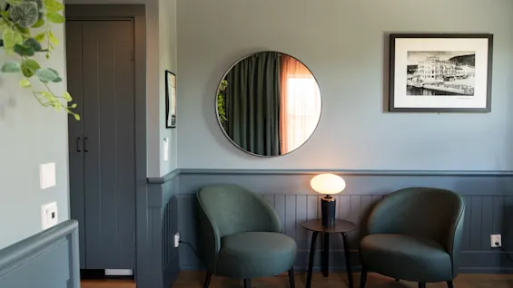 To grønne stole står ved siden af et rundt spejl og et lille bord med en lampe i et gråt værelse. Et indrammet sort-hvidt fotografi hænger på væggen.