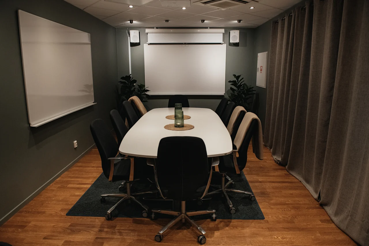 Ett modernt konferensrum med ett stort vitt bord, svarta stolar, whiteboards och en projektorduk, redo för ett möte.
