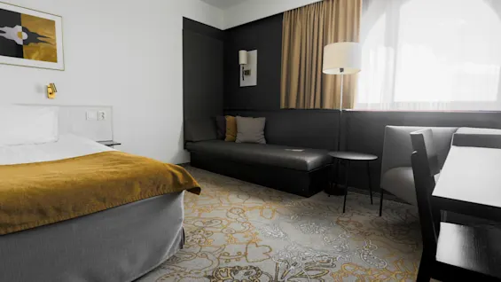 Bed covered with yellow blanket; adjacent sofa in modern hotel room. Soft lighting from lamp near window illuminates patterned carpet and artwork on wall.