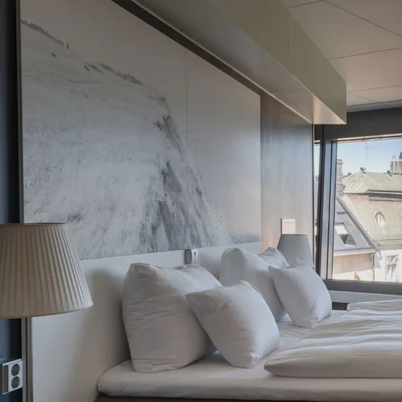 A modern hotel bedroom with a comfortable bed with white linens, bedside lamps, and a snowy mountain picture.