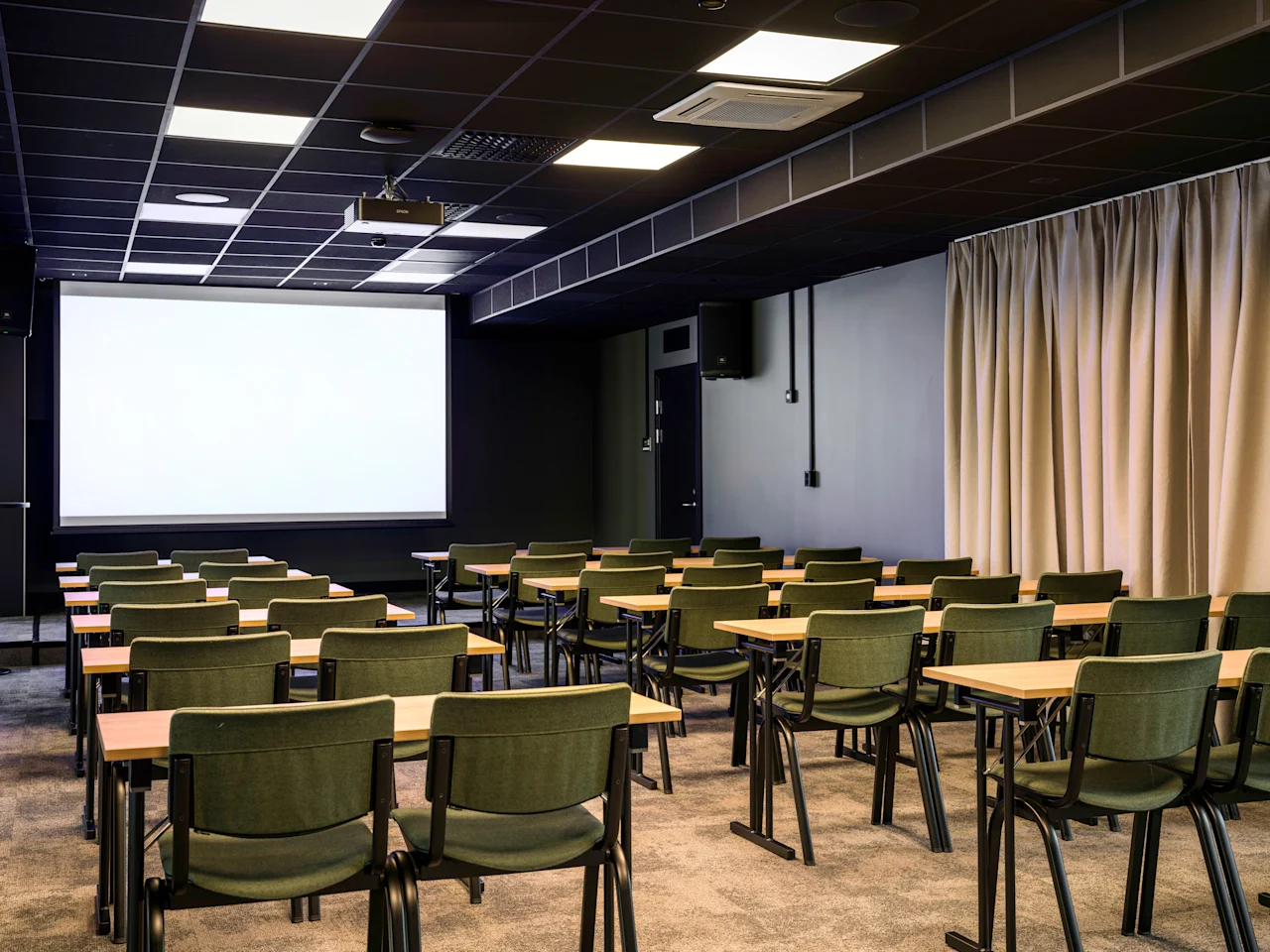A modern conference room with rows of tables and chairs facing a large projection screen, ready for a presentation.