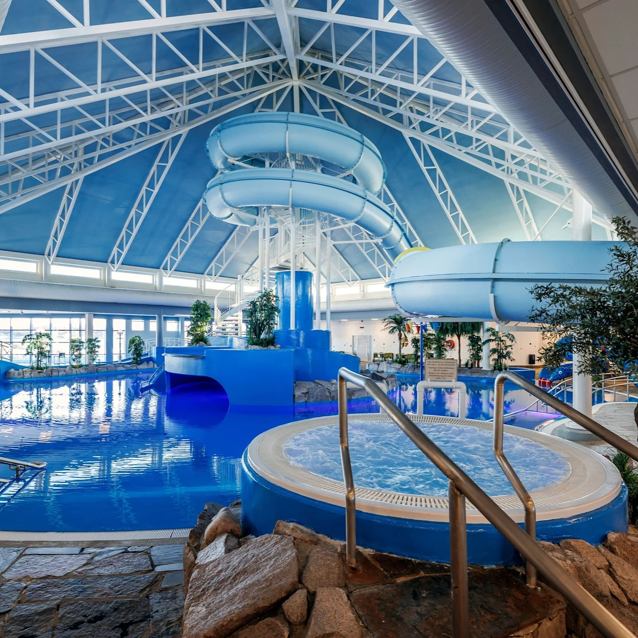 Indoor water park with a winding blue slide above a large pool. Surrounding the area are plants and stone pathways under a high, triangular glass ceiling.