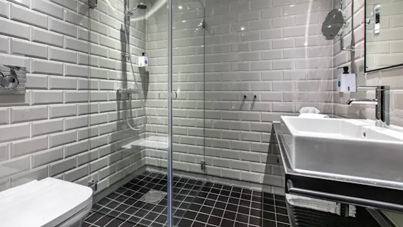 A modern bathroom has beige brick-patterned tiles. A glass shower features a silver showerhead. On the right, a rectangular white sink sits under a mirror with a magnifying extension.