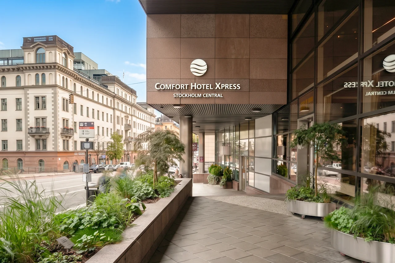 The entrance of a modern hotel in a city with green plants and a classic building in the background.