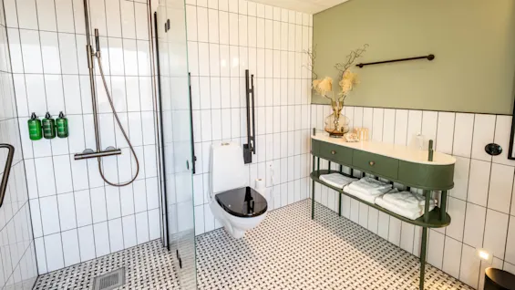 Shower with overhead fixture and glass door. Bathroom features white tiles, green decor items, and a green console table with towels below. Bottles mounted on the wall contain green liquid.