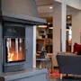 En peis med synlig flamme varmer en koselig stue. I nærheten står en grå lenestol med røde puter på et mønstret teppe, som skaper en avslappende atmosfære.