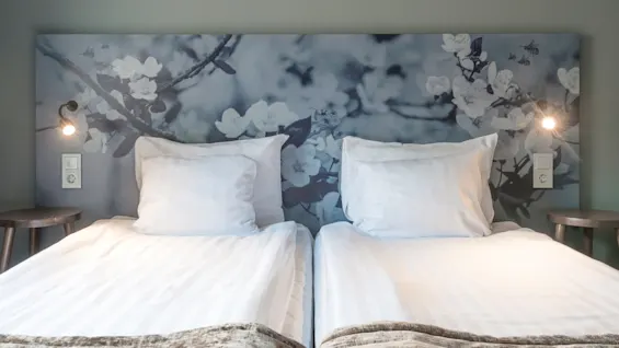 Två prydligt bäddade sängar med vita lakan, huvudgavel dekorerad med blomstermotiv, i ett lugnt sovrum. Två vägglampor ger mjukt ljus på vardera sidan.