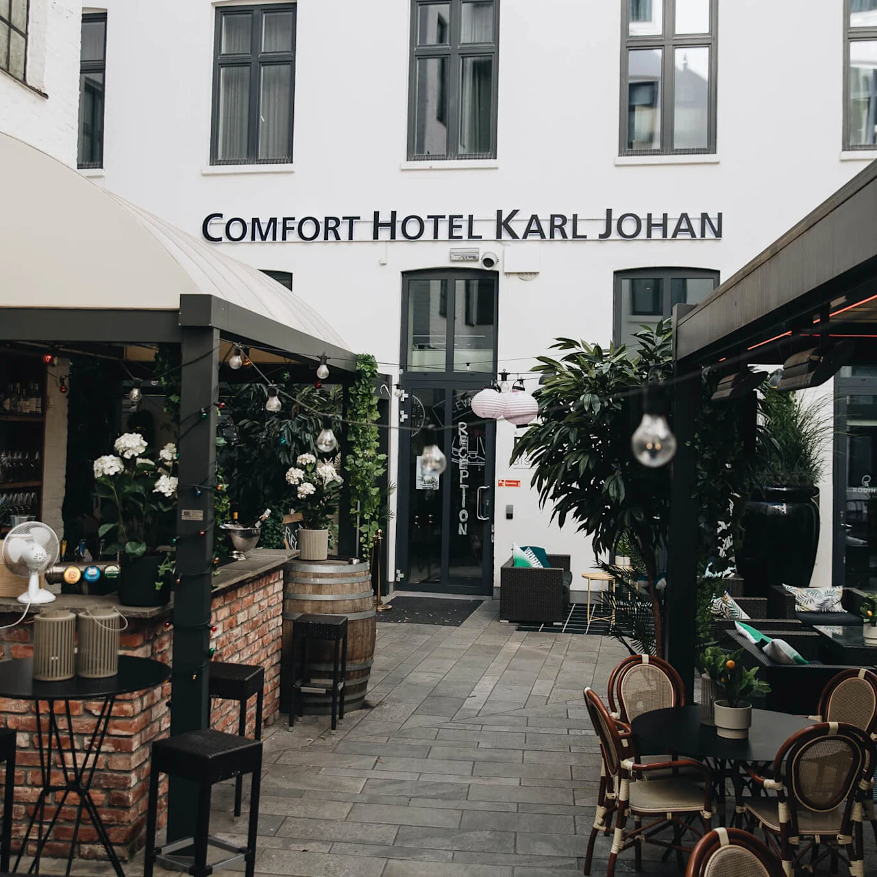 En gårdsplass med bord og stoler omgitt av planter leder til en hotellinngang merket "Comfort Hotel Karl Johan" og "Reception."