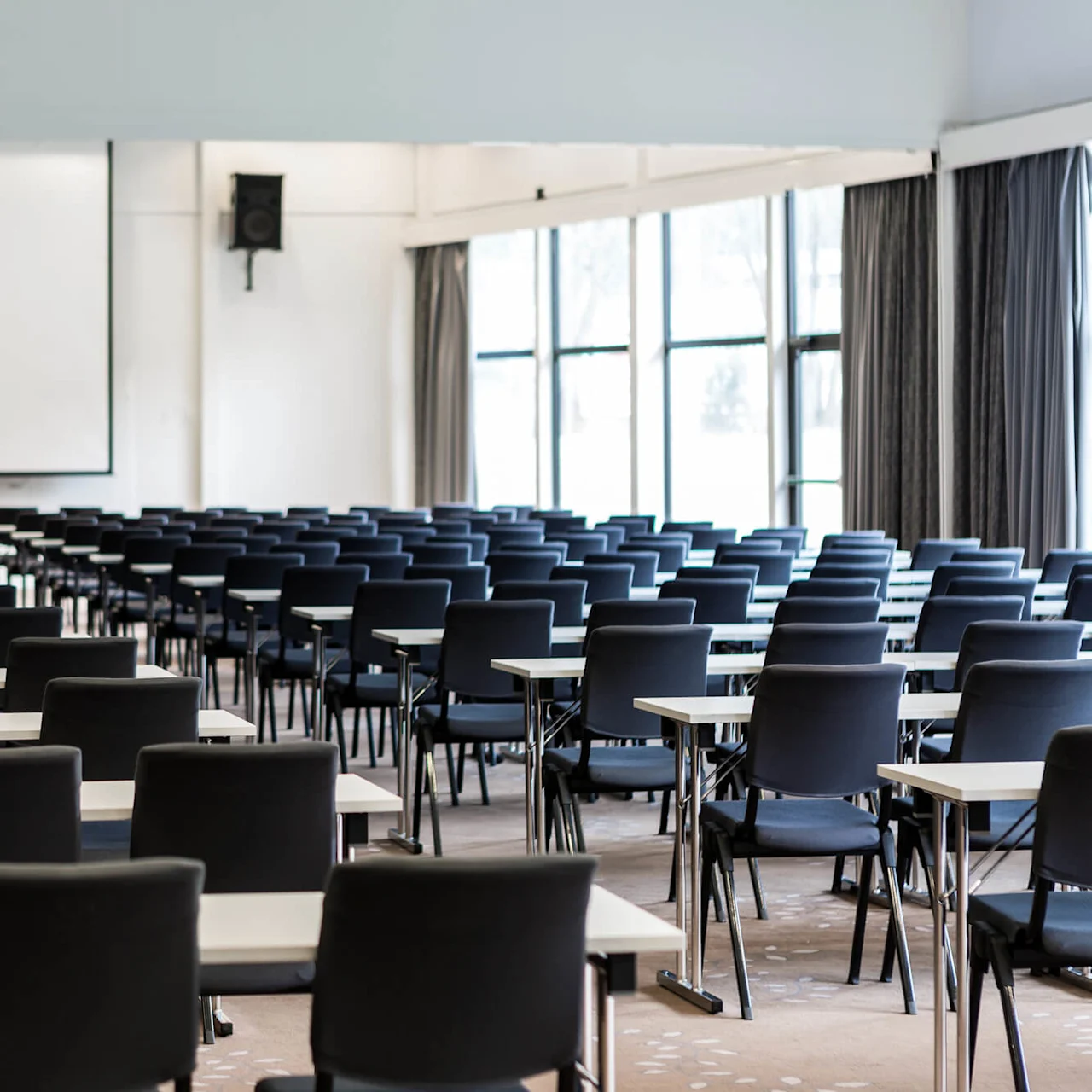 Rows of empty black chairs and white desks face a large projection screen in a spacious conference room with windows and drawn curtains on the side.