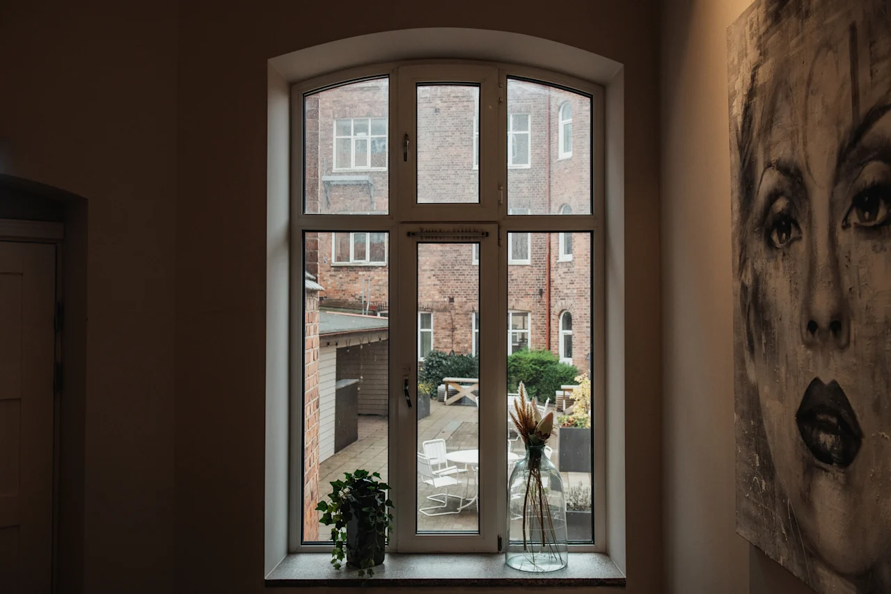 Et vindue vender ud mod en gårdhave med udendørs siddepladser og murstensbygninger. En plante og vase står på vindueskarmen, med kunstværk i nærheden.