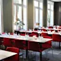 Dining tables with neatly arranged red chairs, set with water bottles and glasses, in a bright room with large windows and gray curtains.