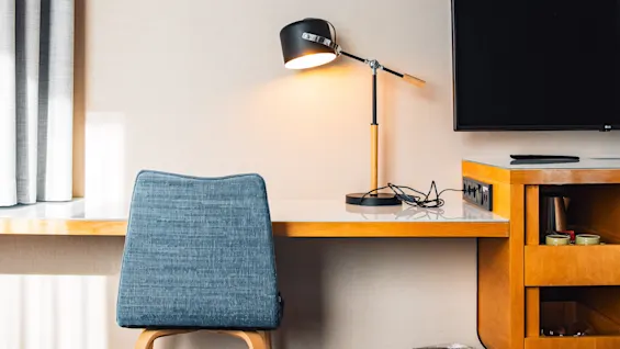 A desk setup with a grey chair and lit desk lamp, adjacent to a TV on the wall, in a hotel room. A trash bin sits underneath.