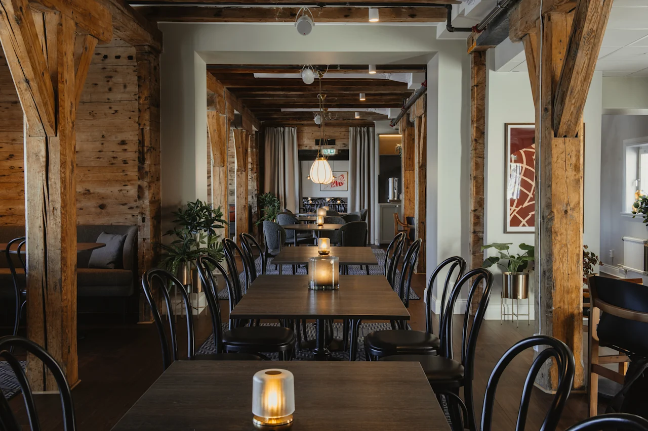 Et hyggeligt restaurantinteriør med træbjælker, dækkede borde og varm belysning, der skaber en indbydende atmosfære til spisning.