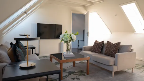 En lys og innbydende stue med komfortable sofaer, et salongbord med blomster og en TV, under skråtak.