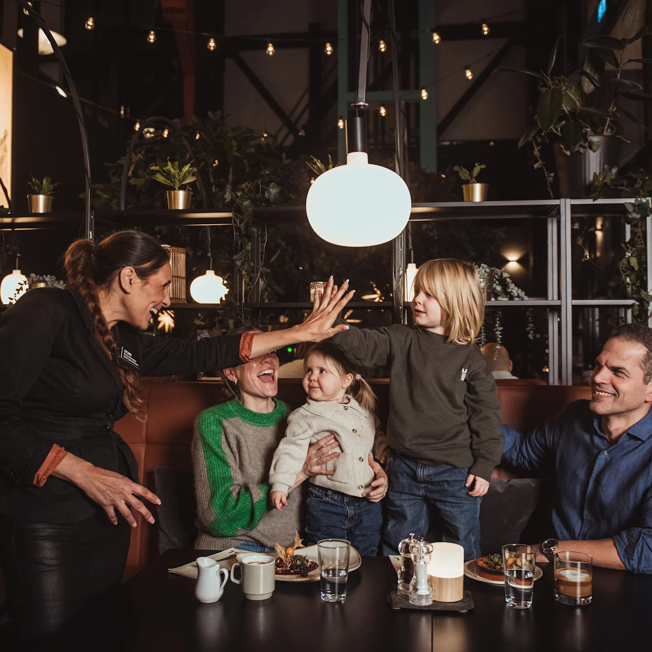 A smiling woman high-fives a young child while others watch, seated around a restaurant table with drinks and plates. Hanging lights and plants adorn the cozy, stylish interior.