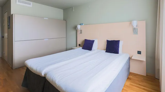 En bäddad dubbelsäng med vita lakan och två lila kuddar står i ett hotellrum, omgiven av ljusa väggar och trägolv.