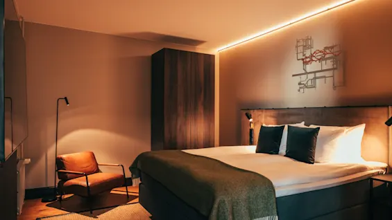 Ett varmt och inbjudande hotellrum med en stor säng, mysig fåtölj och stämningsfull belysning, perfekt för avkoppling.