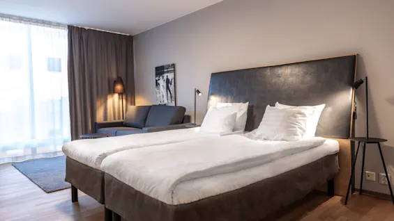 Moderni hotellihuone, jossa kaksi mukavaa sänkyä, viihtyisä sohva ja pehmeä valaistus, valmiina rentouttavaan oleskeluun.
