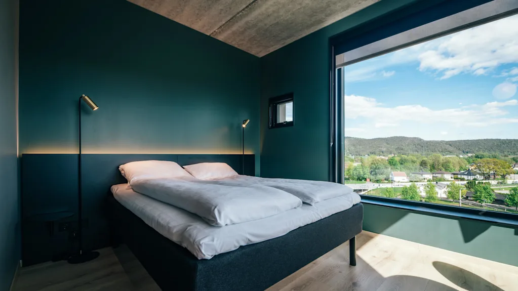 En seng står mot en mørkegrønn vegg, opplyst av en gulvlampe, med et stort vindu som avslører en naturskjønn utsikt over åser og trær.