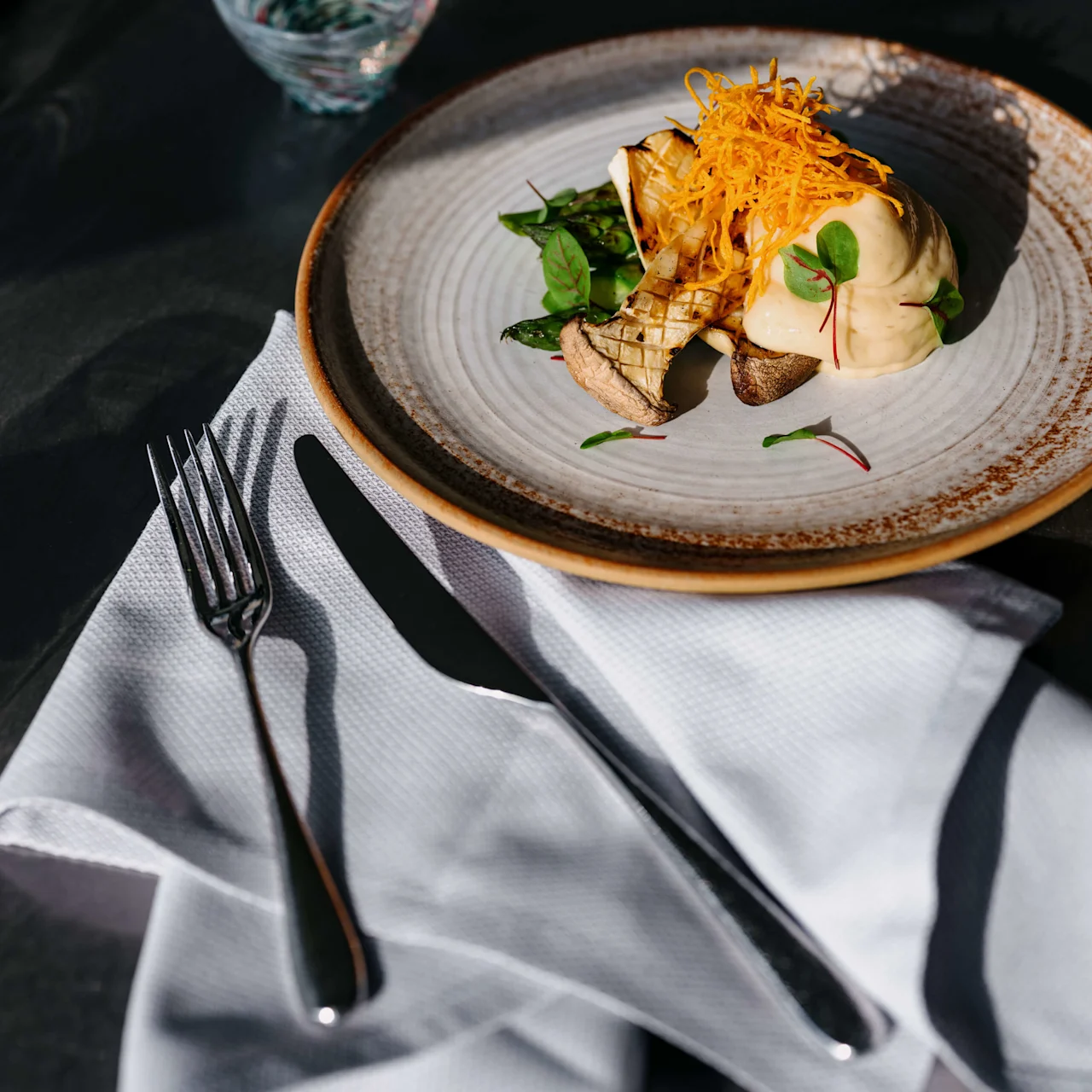 En gourmetret med pynt på en tallerken, sollys kaster skygger, ved siden av bestik på en serviet.