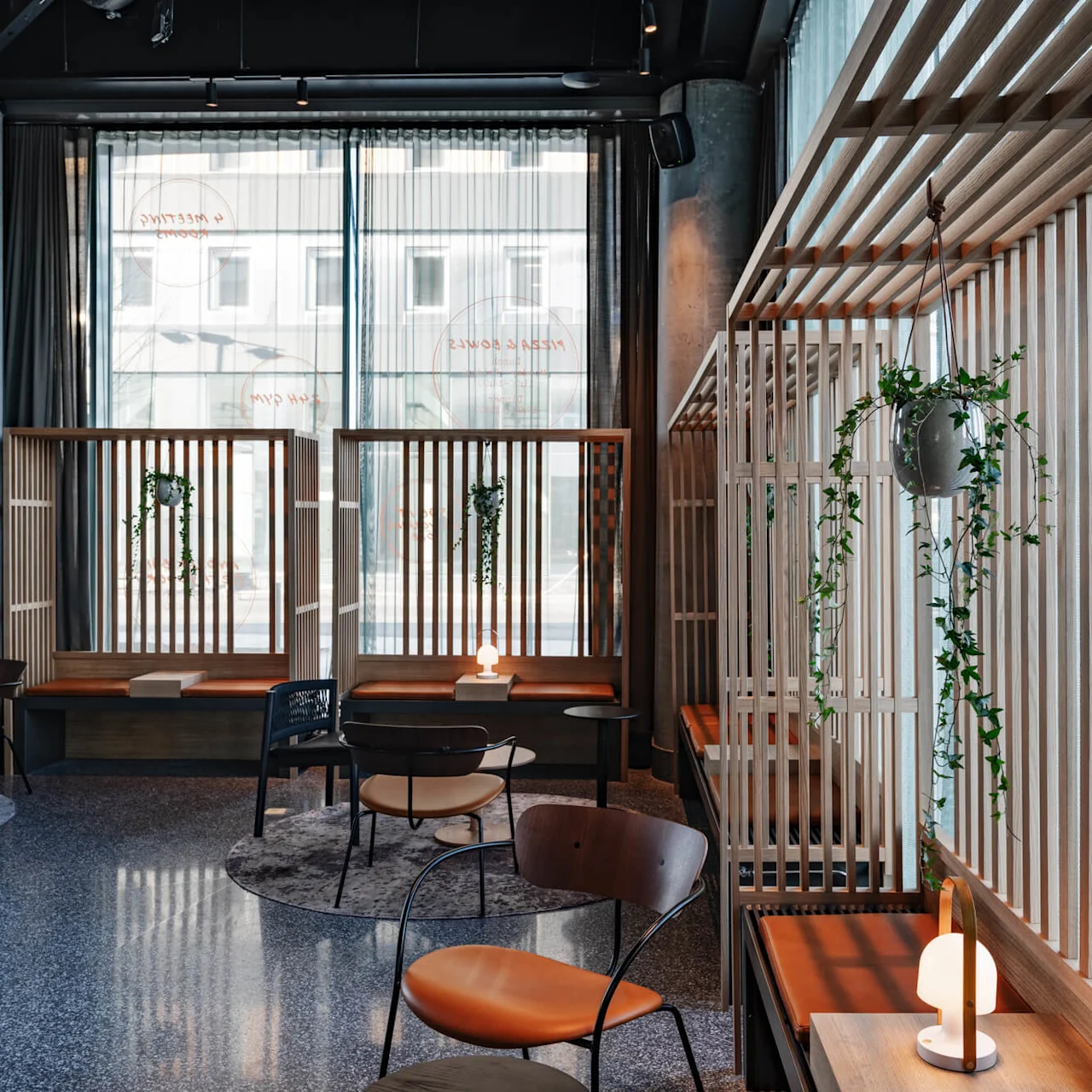 Wooden seating area adorned with small plants and illuminated by a warm lamp; enclosed by wooden slats, within a modern, sunlit interior. Windows display faded text: "MEETING", "RELAX", "BOWLS."