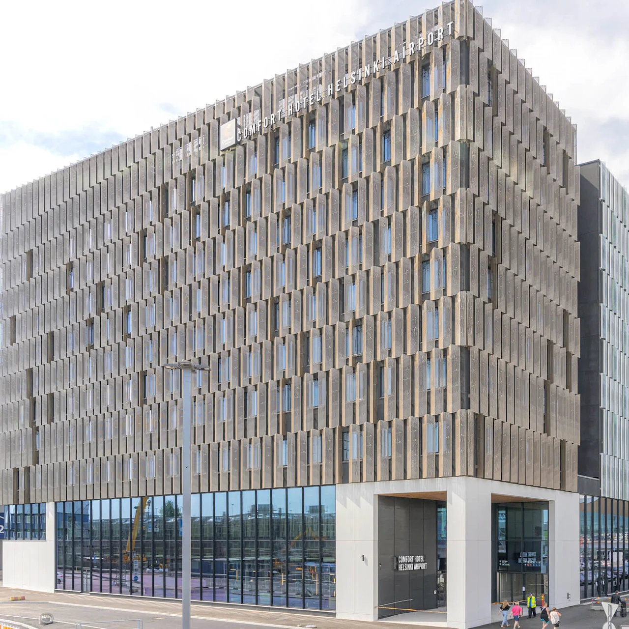 En moderne, geometrisk bygning mærket "COMFORT HOTEL HELSINKI AIRPORT" står nær et parkeringsområde under en delvis skyet himmel. Folk går i nærheden.