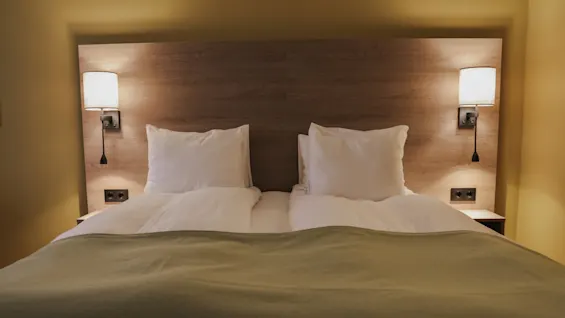 Bed with white pillows and duvet, covered by a green blanket, flanked by two wall lamps on a wooden headboard in a warmly-lit room.