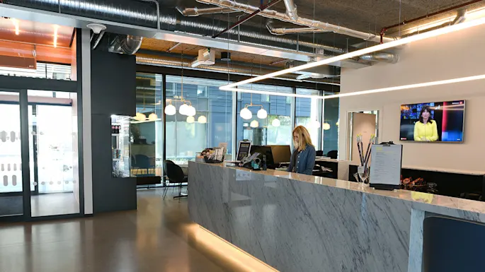 Reception desk with a woman working, surrounded by modern decor and pendant lights. A TV on the wall displays a newscaster. Glass doors lead to a seating area.