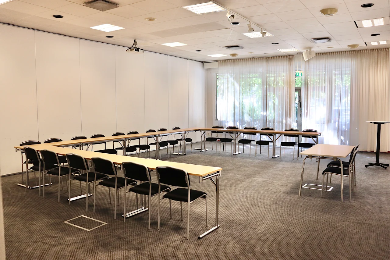 Conference room with U-shaped arrangement of tables and chairs, surrounded by white walls and windows with curtains allowing natural light. Ceiling lights and a projector are visible.
