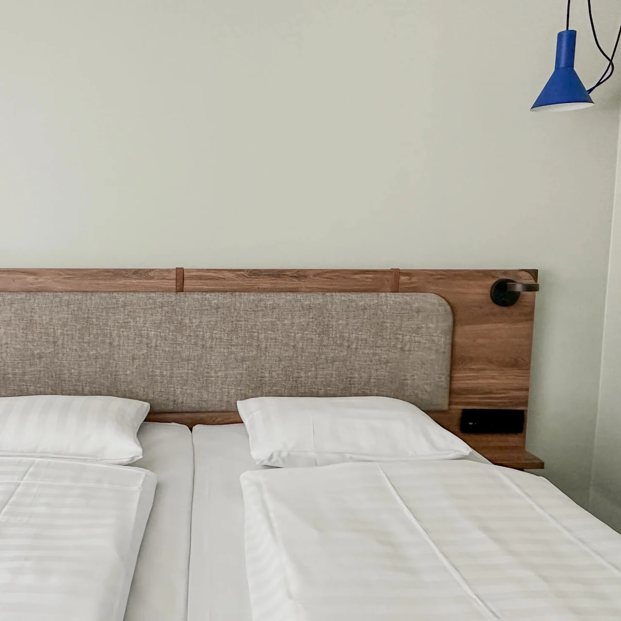 A neatly made hotel bed features white sheets and two pillows, with a wooden headboard and attached lamps, set in a minimalist room with light-colored walls.