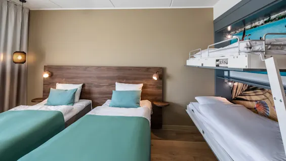 Two single beds with teal bedding are positioned against a wooden headboard, beneath wall lights. A bunk bed with white sheets is on the right. The room features light walls and hardwood flooring.