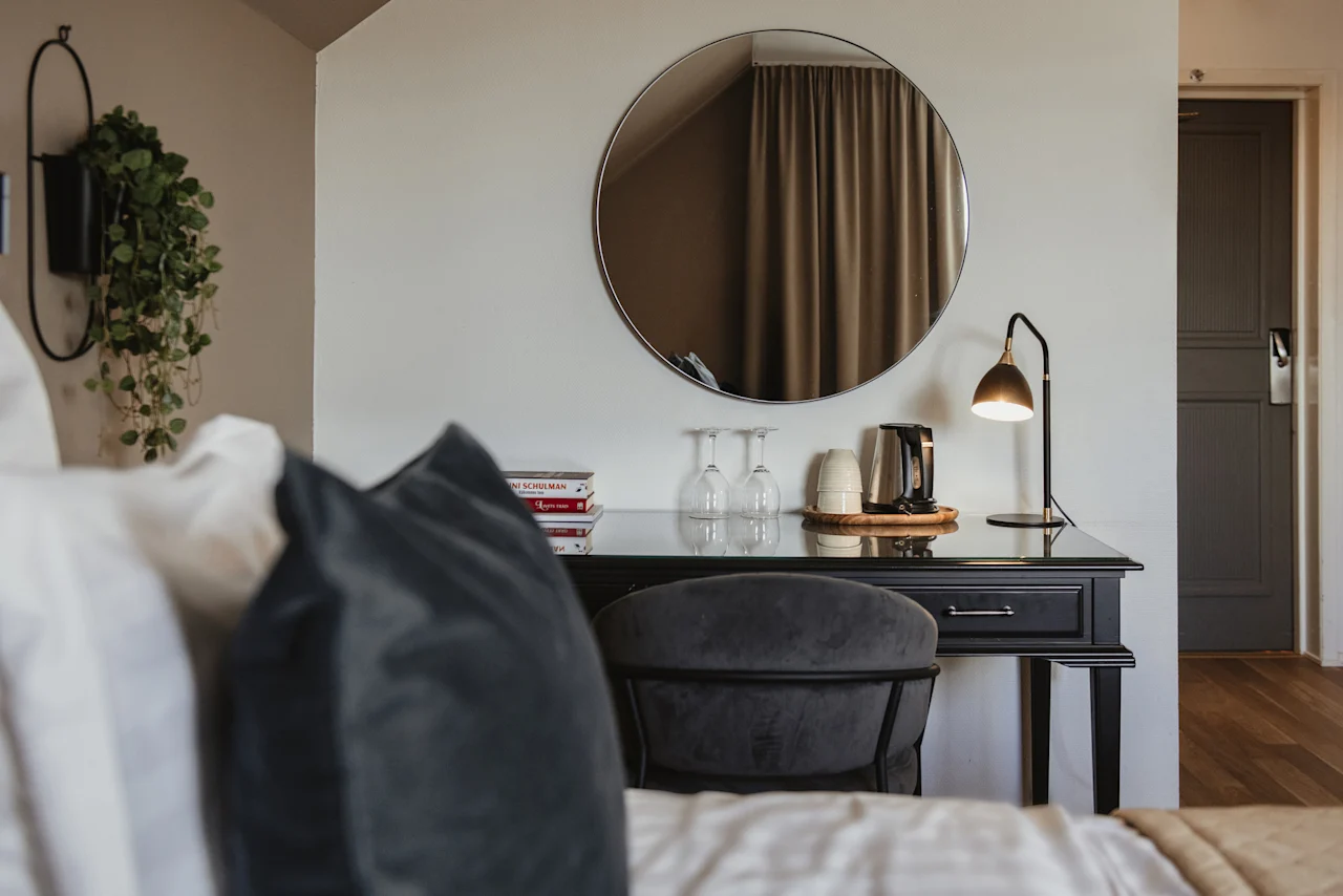 Ett hotellrum med ett skrivbord, stol och en spegel. Böcker N SCHULMAN och Tage, en vattenkokare och glas står på skrivbordet.