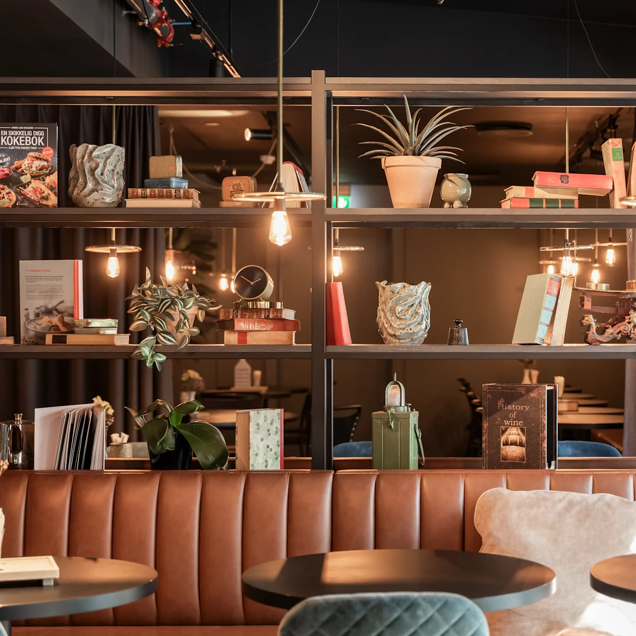 En hyggelig café med brun lædersofa, runde borde og hylder dekoreret med planter og bøger. Varm belysning og små potteplanter skaber en indbydende atmosfære. Synlige bogtitler: "KOKEBOK" og "Kokekatere".