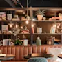 En hyggelig café med brun lædersofa, runde borde og hylder dekoreret med planter og bøger. Varm belysning og små potteplanter skaber en indbydende atmosfære. Synlige bogtitler: "KOKEBOK" og "Kokekatere".