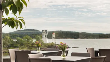 Bord arrangert på en treterrasse har vaser med fargerike blomster; grønne trær og en rolig innsjø med fjerne åser danner den rolige bakgrunnen.
