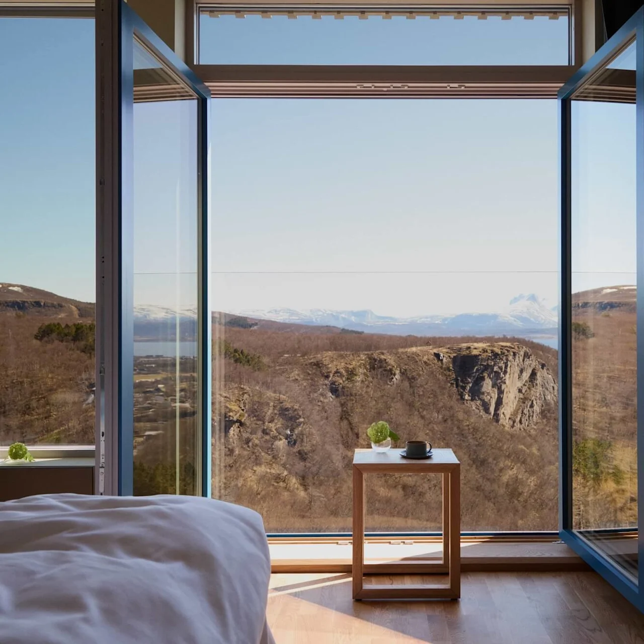 Open window frames a stunning mountain view; a wooden table holds a cup and plant. Inside, part of a bed with white sheets is visible.