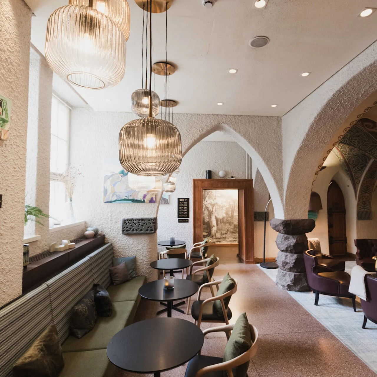 Elegant lounge features cushioned seating and round tables under ornate arches. Warm lighting from hanging lamps enhances the cozy atmosphere. A sign reads "TOILETS."