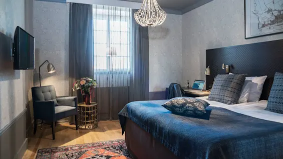 Et pent ordnet hotellrom med en stor seng, lenestol og skrivebord under et vindu med transparente gardiner og en lysekrone.