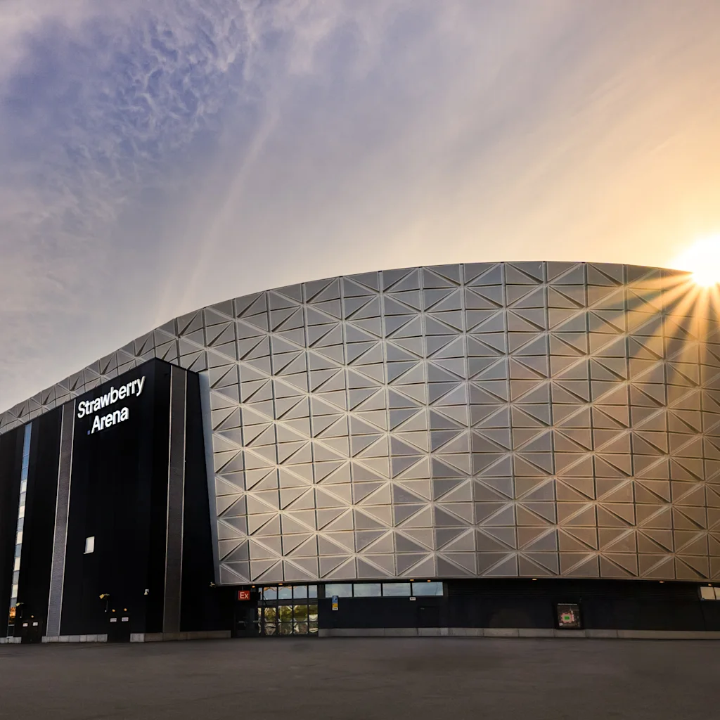 Strawberry Arena-bygningen ved solnedgang, med solen der skinner stærkt bag den. Bygningen har en moderne, geometrisk facade.