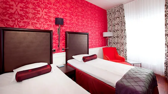 Two twin beds with dark headboards against a vibrant red wallpapered wall, accented by red and white decor, in a stylish bedroom.