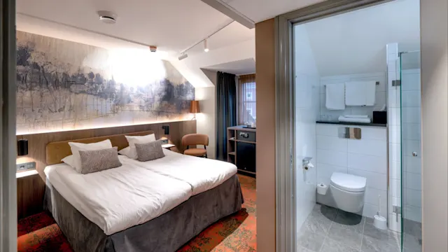 A hotel room with two beds and abstract wall art, adjacent to a bathroom featuring tiled walls and a hanging toilet; a small window provides ambient light.