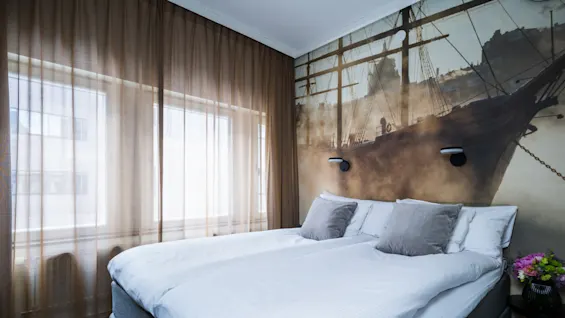 A neatly made bed sits in a room with sheer curtains filtering light, complemented by a ship-themed mural on the wall and a flower vase on a side table.