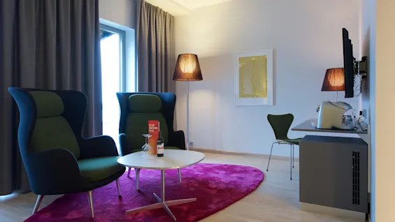 Två gröna fåtöljer står på en rosa matta bredvid ett bord med en vinflaska i ett modernt rum. Text: "GET ME SOMETHING TO EAT."